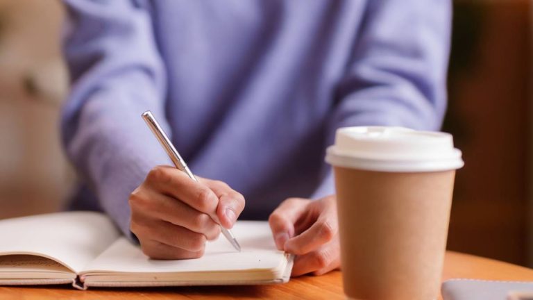 Woman in lavender sweater writing in journal at wooden table with takeout coffee.