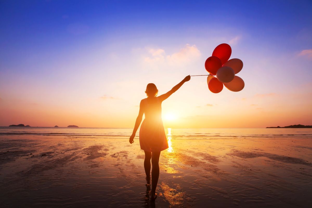 Woman walking into sunset with handful of helium balloons.